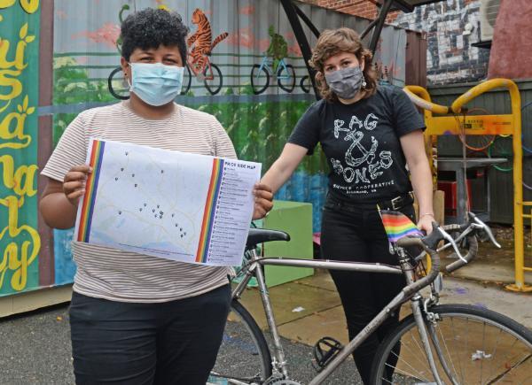 Two people standing. The person on the left holds a Pride Ride map. The person on the right is leaning on a bike.